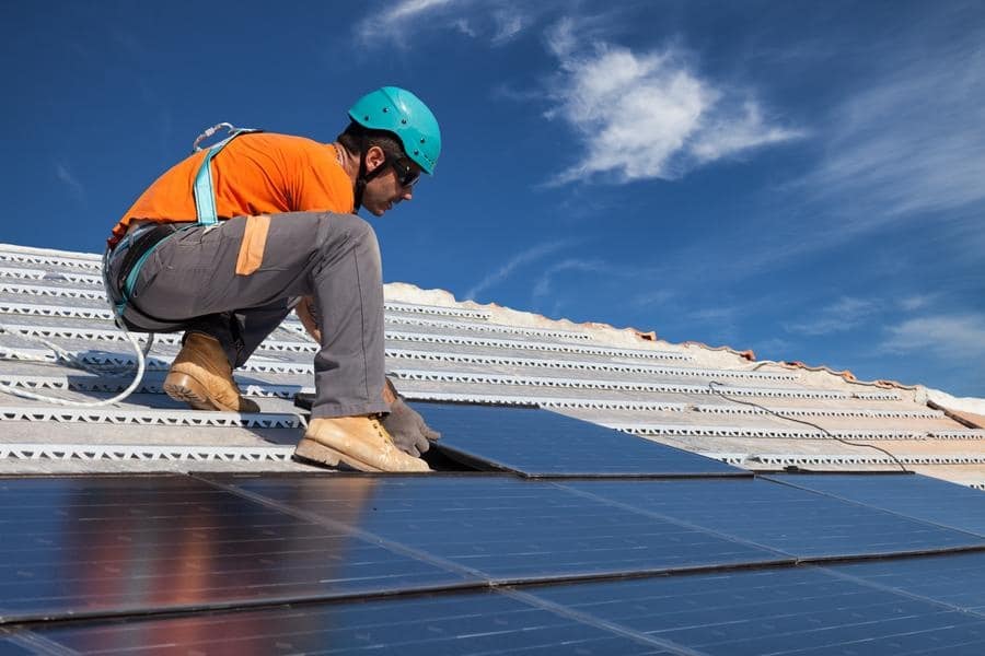 A worker wearing safety gear installs solar panels on a rooftop under a blue sky.