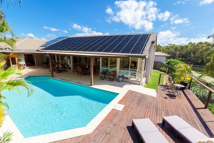 A modern house with a rooftop solar panel array, a swimming pool, lounge chairs on a wooden deck, and outdoor seating areas, surrounded by greenery under a blue sky.