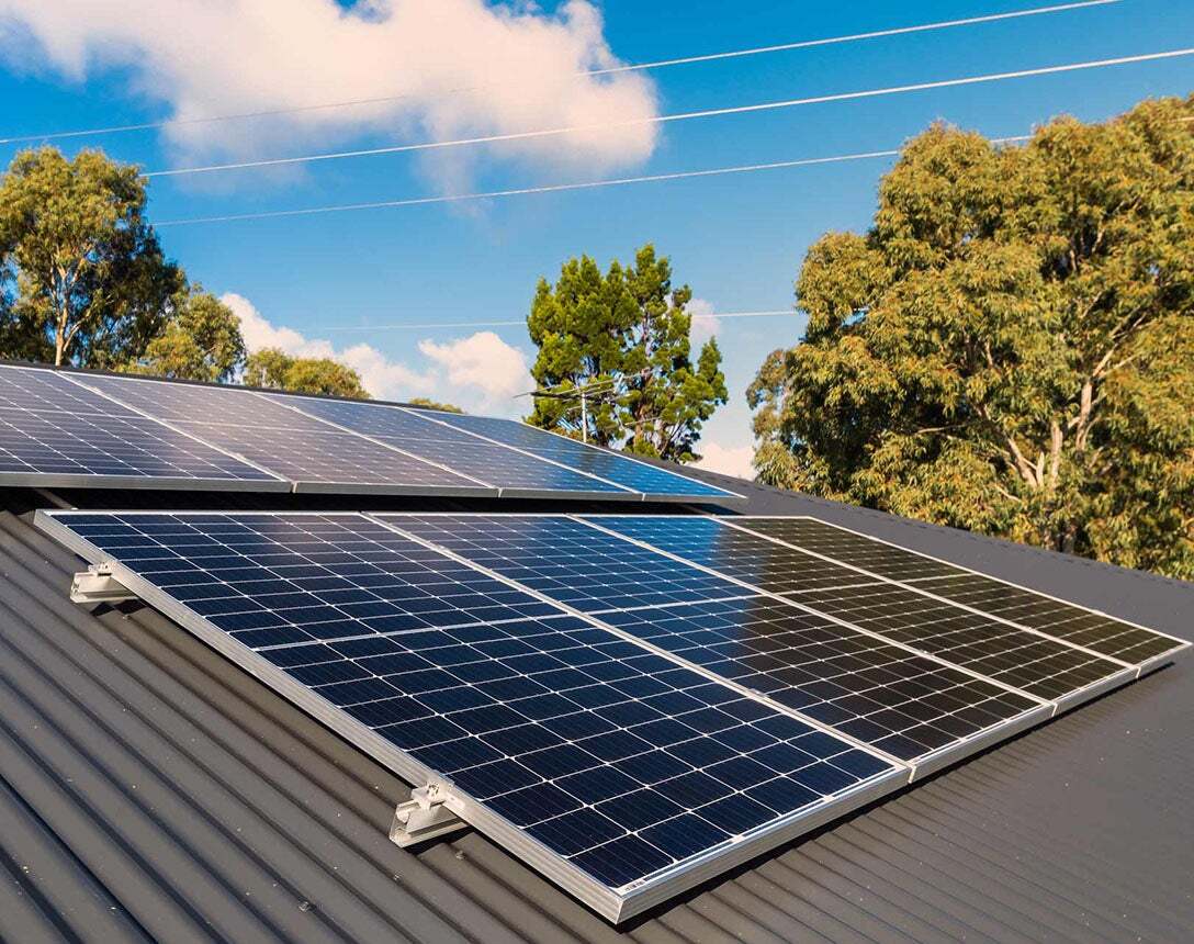 Solar panels installed on a sloped metal roof with trees and power lines in the background under a partly cloudy sky.