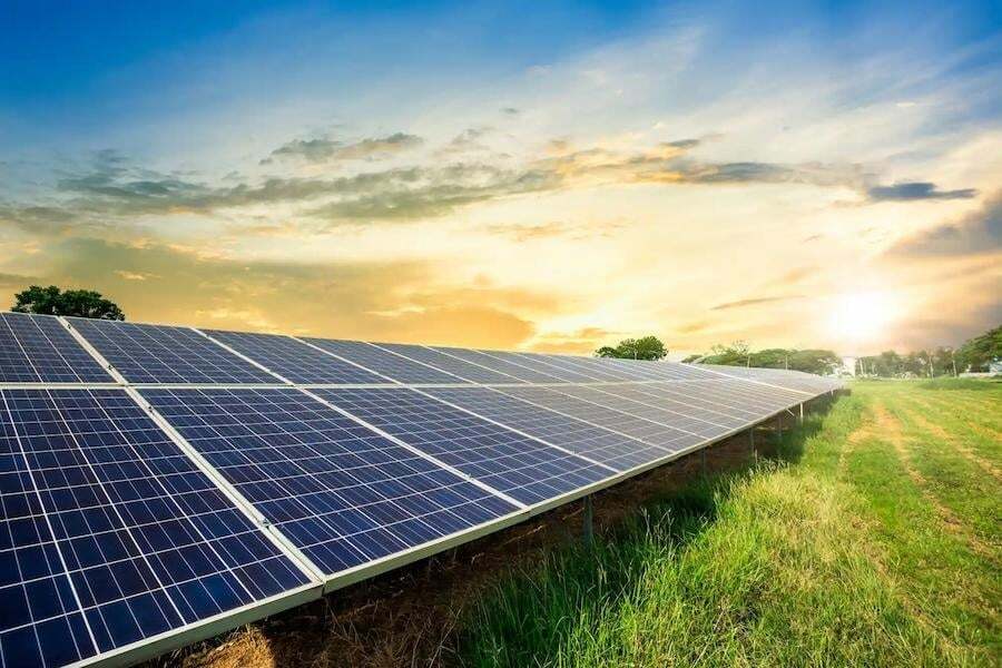 Rows of solar panels installed on a grassy field with the sun setting in the background and a partly cloudy sky above.