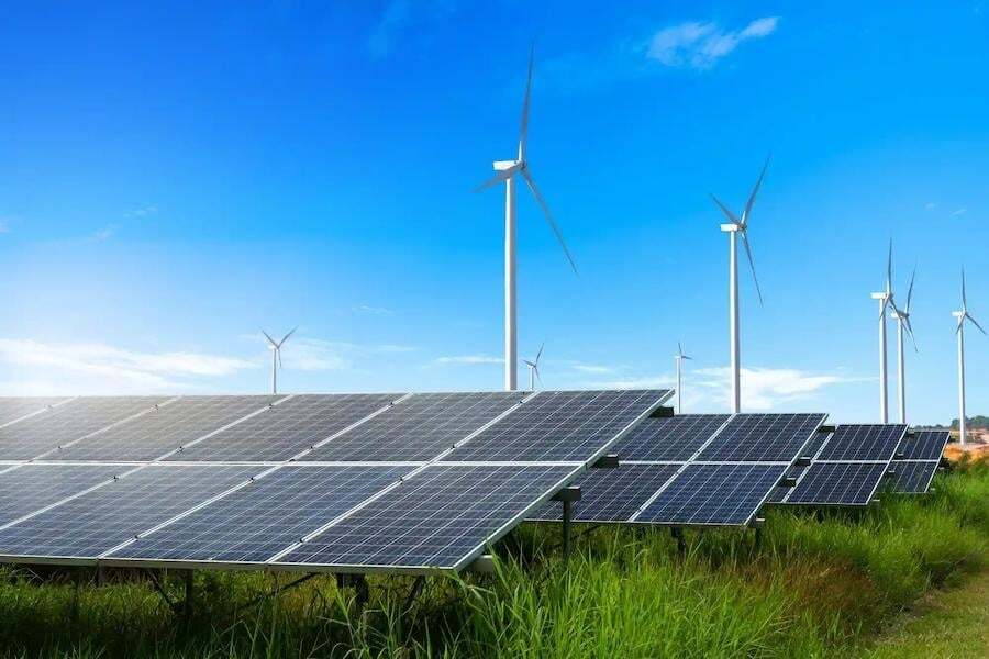 Rows of solar panels in the foreground with wind turbines in the background, set in a grassy field under a clear blue sky.