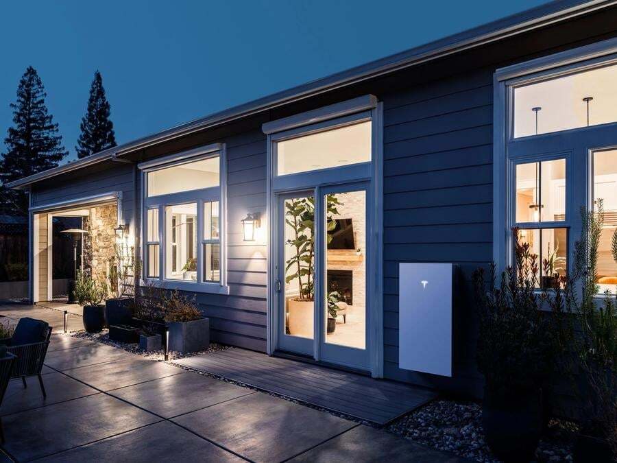 Modern house exterior at dusk with large windows illuminated from inside and a Tesla Powerwall battery mounted on the outside wall.